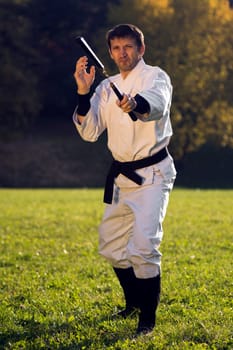 Ninja in white kimono is practicing with nunchaku outdoors in the park.