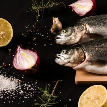 Raw trout fish with salt, pepper, lemon, garlic, onion and rosemary on black background.