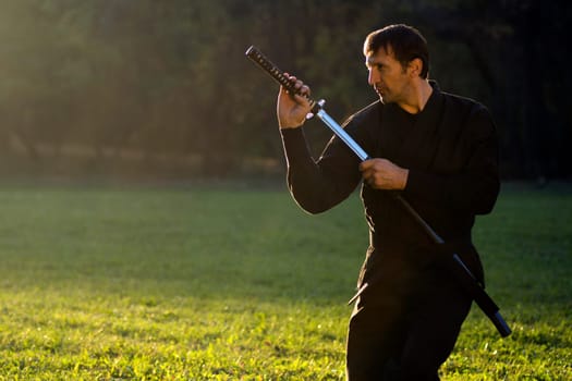 Ninja in black kimono with sword is outdoors in the park.