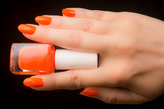 Female hand with fingers holds orange nail polish on black background, orange colored manicure.