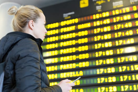 Young woman at international airport looking at the flight information board, holding smart phone in her hand, checking flight informations on phone application.