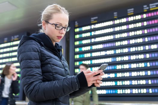 Woman in international airport looking at smart phone app information and flight information board, checking her flight detailes.