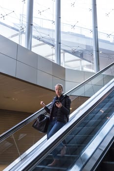 Young businesswoman with large black bag and mobile phone getting down on the escalator during business trip in modern city.