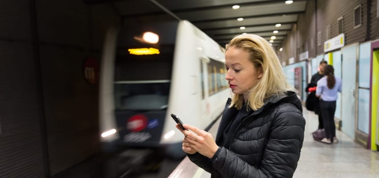 Young casual woman using mobile phone in her hand waiting on the platform of a metro station for metro to arrive. Public transport.