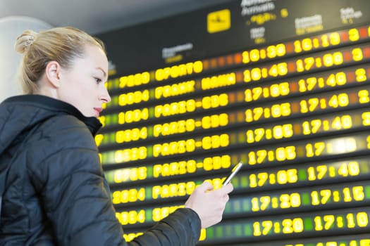 Young woman at international airport looking at the flight information board, holding smart phone in her hand, checking flight informations on phone application.