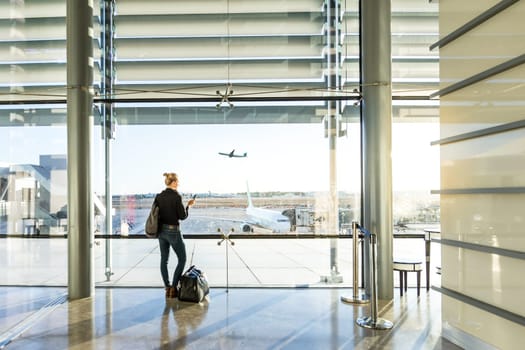 Young casual female traveler at airport, holding smart phone device, looking through the airport gate windows at planes on airport runway.