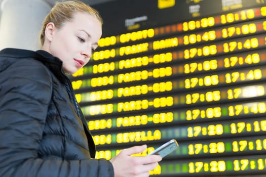 Young woman at international airport looking at the flight information board, holding smart phone in her hand, checking flight informations on phone application.
