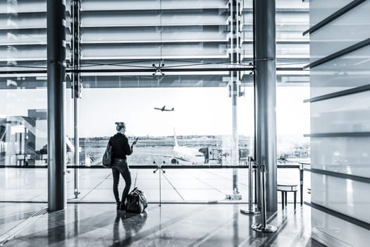 Young casual female traveler at airport, holding smart phone device, looking through the airport gate windows at planes on airport runway.