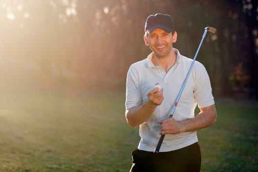 Portrait of handsome smiling golfer with golf club and ball outdoors on playing field.