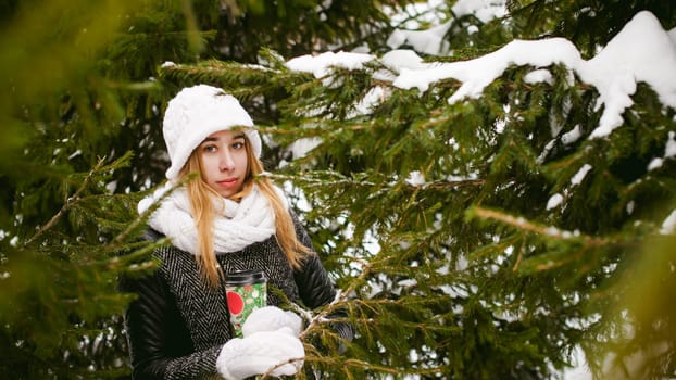 Portrait of cute woman in white scarf and hat knitted coat on outdoors background of snow and blurred fir branches in winter. girl holds a coffee cup in hand, christmas takeaway coffee