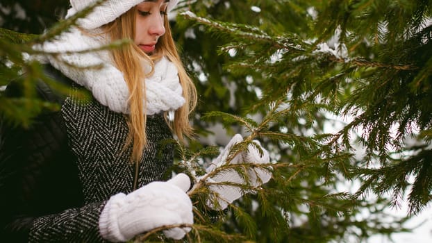 young woman in a forest with fir and pine trees on the street, hands touch the branches of trees, considering them. peridium winter, warm clothes, knitted mittens and scarf white