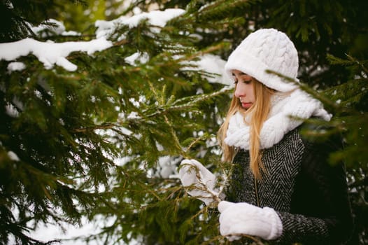 young woman in a forest with fir and pine trees on the street, hands touch the branches of trees, considering them. peridium winter, warm clothes, knitted mittens and scarf white