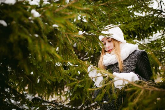 young woman in a forest with fir and pine trees on the street, hands touch the branches of trees, considering them. peridium winter, warm clothes, knitted mittens and scarf white