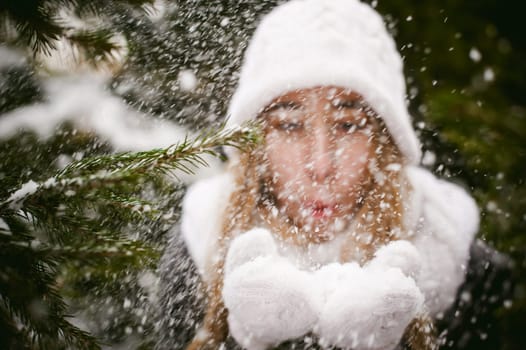 Blowing Snow. Beautiful Joyful Female Model on background of fir trees. Young woman enjoying magical snowfall. Winter wonderland.