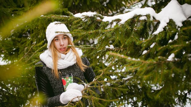 Portrait of cute woman in white scarf and hat knitted coat on outdoors background of snow and blurred fir branches in winter. girl holds a coffee cup in hand, christmas takeaway coffee