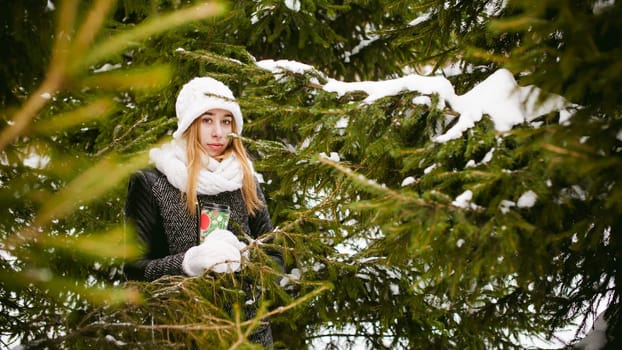 Portrait of cute woman in white scarf and hat knitted coat on outdoors background of snow and blurred fir branches in winter. girl holds a coffee cup in hand, christmas takeaway coffee