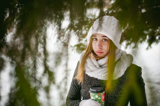 Portrait of cute woman in white scarf and hat knitted coat on outdoors background of snow and blurred fir branches in winter. girl holds a coffee cup in hand, christmas takeaway coffee