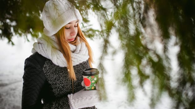 Portrait of cute woman in white scarf and hat knitted coat on outdoors background of snow and blurred fir branches in winter. girl holds a coffee cup in hand, christmas takeaway coffee
