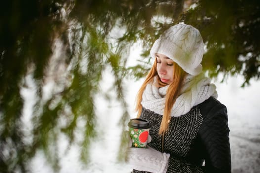 Portrait of cute woman in white scarf and hat knitted coat on outdoors background of snow and blurred fir branches in winter. girl holds a coffee cup in hand, christmas takeaway coffee