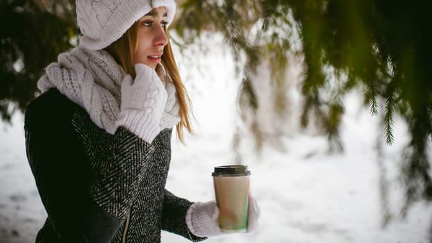 Portrait of cute woman in white scarf and hat knitted coat on outdoors background of snow and blurred fir branches in winter. girl holds a coffee cup in hand, christmas takeaway coffee