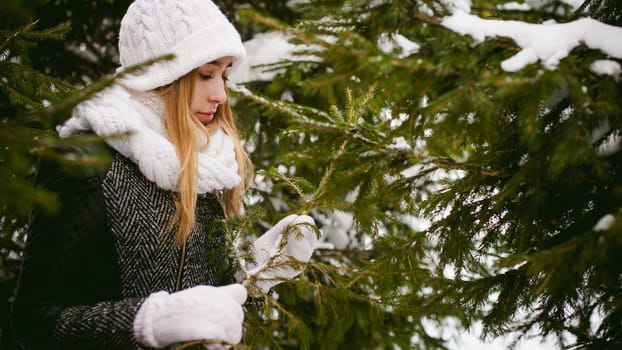young woman in a forest with fir and pine trees on the street, hands touch the branches of trees, considering them. peridium winter, warm clothes, knitted mittens and scarf white