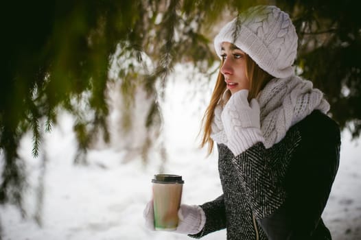 Portrait of cute woman in white scarf and hat knitted coat on outdoors background of snow and blurred fir branches in winter. girl holds a coffee cup in hand, christmas takeaway coffee