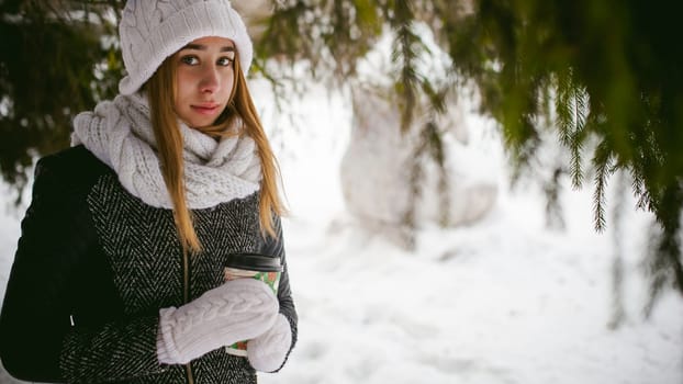 Portrait of cute woman in white scarf and hat knitted coat on outdoors background of snow and blurred fir branches in winter. girl holds a coffee cup in hand, christmas takeaway coffee