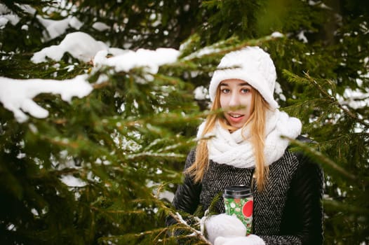 Portrait of cute woman in white scarf and hat knitted coat on outdoors background of snow and blurred fir branches in winter. girl holds a coffee cup in hand, christmas takeaway coffee