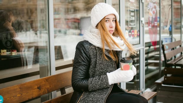 woman in white scarf and hat drinks coffee. cute young blonde girl sitting on a bench near the coffee shop, drinking takeaway coffee, holding Christmas paper cup in his hands. looking at the camera
