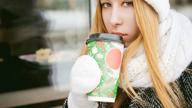 woman in white scarf and hat drinks coffee. cute young blonde girl sitting on a bench near the coffee shop, drinking takeaway coffee, holding Christmas paper cup in his hands. looking at the camera