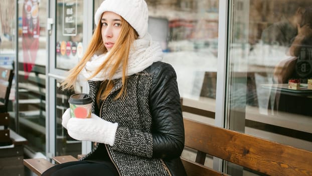woman in white scarf and hat drinks coffee. cute young blonde girl sitting on a bench near the coffee shop, drinking takeaway coffee, holding Christmas paper cup in his hands. looking at the camera