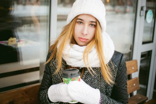 woman in white scarf and hat drinks coffee. cute young blonde girl sitting on a bench near the coffee shop, drinking takeaway coffee, holding Christmas paper cup in his hands. looking at the camera