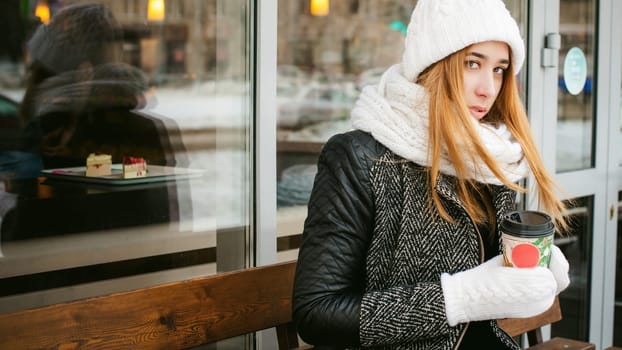woman in white scarf and hat drinks coffee. cute young blonde girl sitting on a bench near the coffee shop, drinking takeaway coffee, holding Christmas paper cup in his hands. looking at the camera