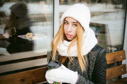 woman in white scarf and hat drinks coffee. cute young blonde girl sitting on a bench near the coffee shop, drinking takeaway coffee, holding Christmas paper cup in his hands. looking at the camera