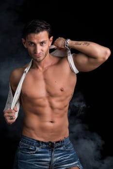 Handsome muscular man pulling up t-shirt on naked torso, on dark background