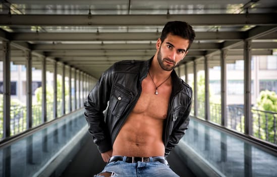 Handsome bearded young man sitting, wearing leather jacket on naked torso, outdoors in urban environment