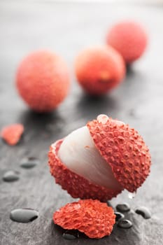 Four lychee fruits on slate plate. Front fruit with open peel and a piece of peel with water drops on slate plate in front of it in focus. Rear fruits fuzzy.