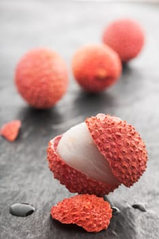 Four lychee fruits on slate plate. Front fruit with open peel and a piece of peel with water drops on slate plate in front of it in focus. Rear fruits fuzzy.