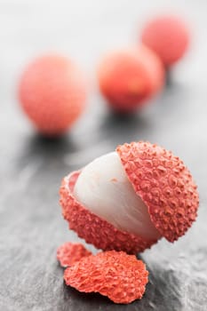 Four lychee fruits on slate plate. Front fruit with open peel and a piece of peel in front of it on slate plate in focus. Rear fruits blurred as a bright silhouette.