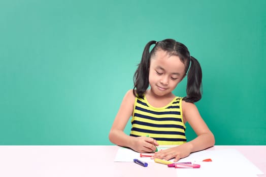Cute asian little girl drawing with crayon while sitting at table over green background