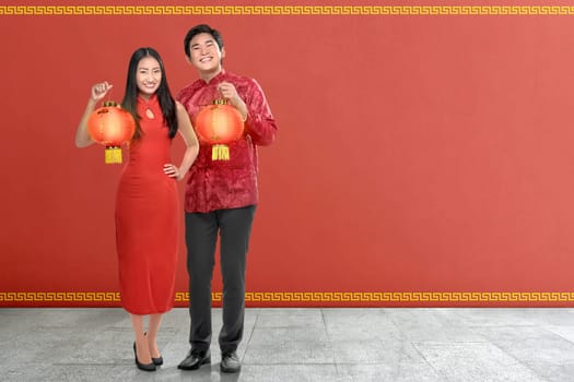 Young chinese couple with traditional dress holding red lanterns on red wall background. Happy Chinese New Year