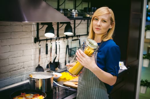 woman housewife preparing in the kitchen. Smiling young blond woman in an apron, holding a glass jar with spaghetti, standing at the stove