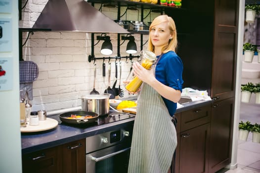 woman housewife preparing in the kitchen. Smiling young blond woman in an apron, holding a glass jar with spaghetti, standing at the stove
