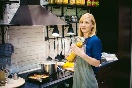 woman housewife preparing in the kitchen. Smiling young blond woman in an apron, holding a glass jar with spaghetti, standing at the stove