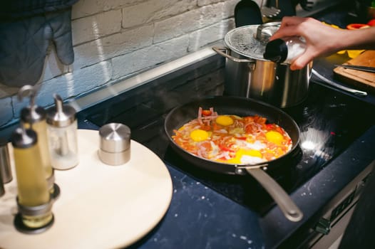 woman housewife prepares fried eggs with ham and vegetables in his kitchen. egg breaks, adding to the pan