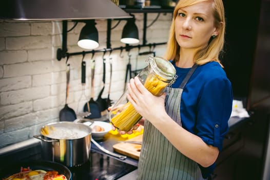 woman housewife preparing in the kitchen. Smiling young blond woman in an apron, holding a glass jar with spaghetti, standing at the stove