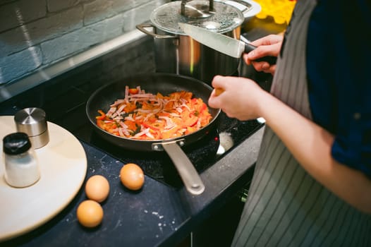 woman housewife prepares fried eggs with ham and vegetables in his kitchen. egg breaks, adding to the pan