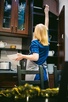 woman housewife in the kitchen. girl pulls out from the shelf cupboard dishes for table service. against the background of the Christmas tree