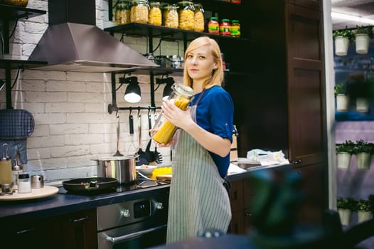 woman housewife preparing in the kitchen. Smiling young blond woman in an apron, holding a glass jar with spaghetti, standing at the stove
