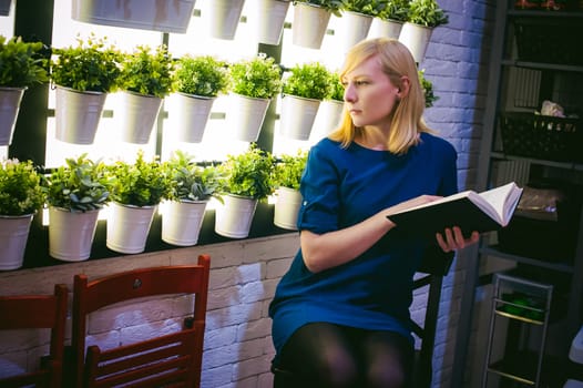 blonde woman in a dress sits on a chair with a book in hand, against the backdrop of houseplants. engaged in reading